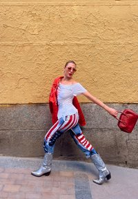 Bela srajca brez naramnic, rdeča jakna, modri kavbojki z vzorcem ameriške zastave, srebrni kavbojski čevlji in rdeča torbica, postavljeni ob rumeno steno.
