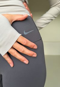 Gros plan sur un legging gris Nike avec une texture lisse, arborant un logo Nike gris clair, et une main reposant sur le tissu. Haut à manches longues visible.