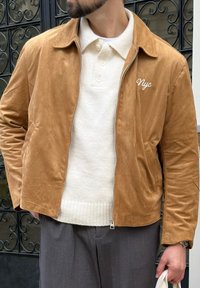 Egy férfi bézs velúr dzsekit visel, amelyen "Nyc" hímzés látható, krémszínű kötött pólóval, szürke nadrággal, és egy fehér vászontáskával áll egy fekete kovácsoltvas kapu mellett.