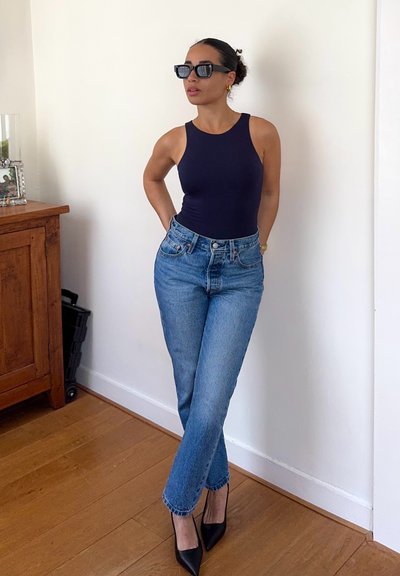 Mujer con top sin mangas azul oscuro, jeans azul claro, tacones negros y gafas de sol, de pie con las manos detrás de la espalda cerca de un armario de madera.