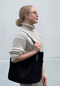 Blondinka z očali v bež pletenem puloverju z visokim ovratnikom drži veliko črno zamšasto torbo, stoji pred enobarvno svetlo steno.