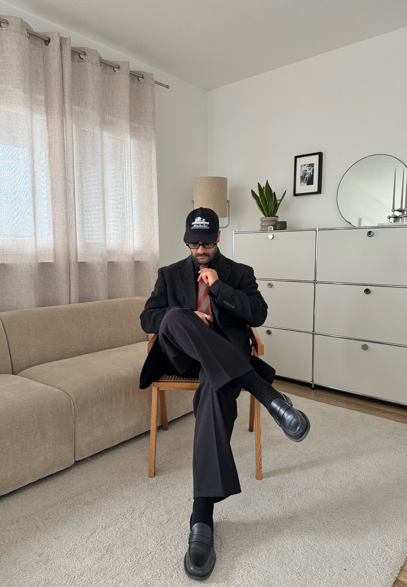 Man wearing black suit, striped tie, and cap sits cross-legged on wooden chair in modern living room with beige sofa and white cabinet.