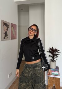 Jeune personne aux cheveux roux et portant de grandes lunettes noires, vêtue d'une veste zippée Adidas noire et d'un pantalon camouflage, se tenant à l'intérieur près d'une œuvre murale et d'une plante.