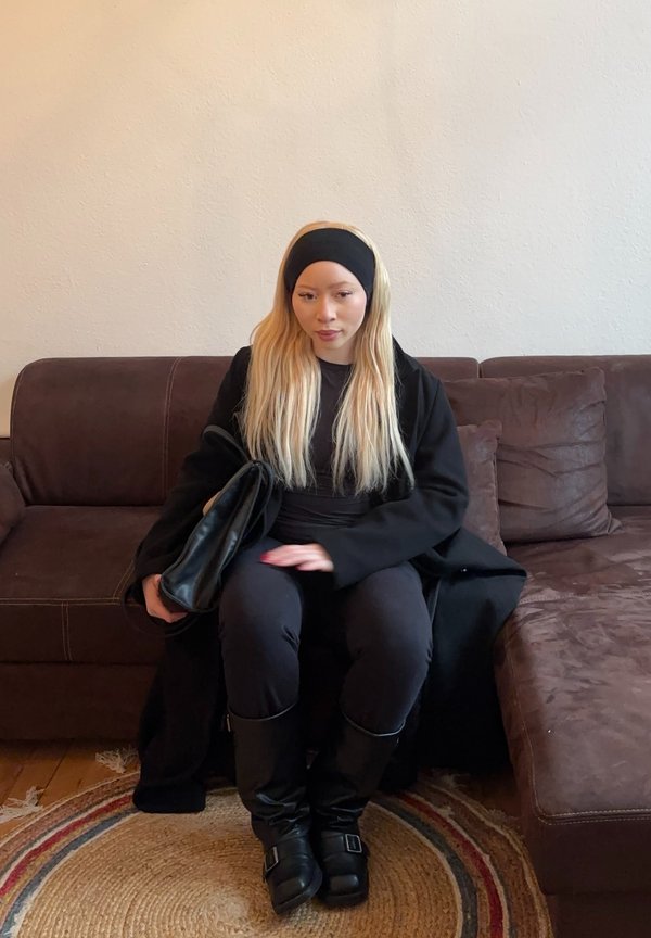 Zwarte jas en top, zwarte broek en zwarte laarzen tot de knie. Lang blond haar; zittend op een donkerbruine bank met een ronde tapijt.