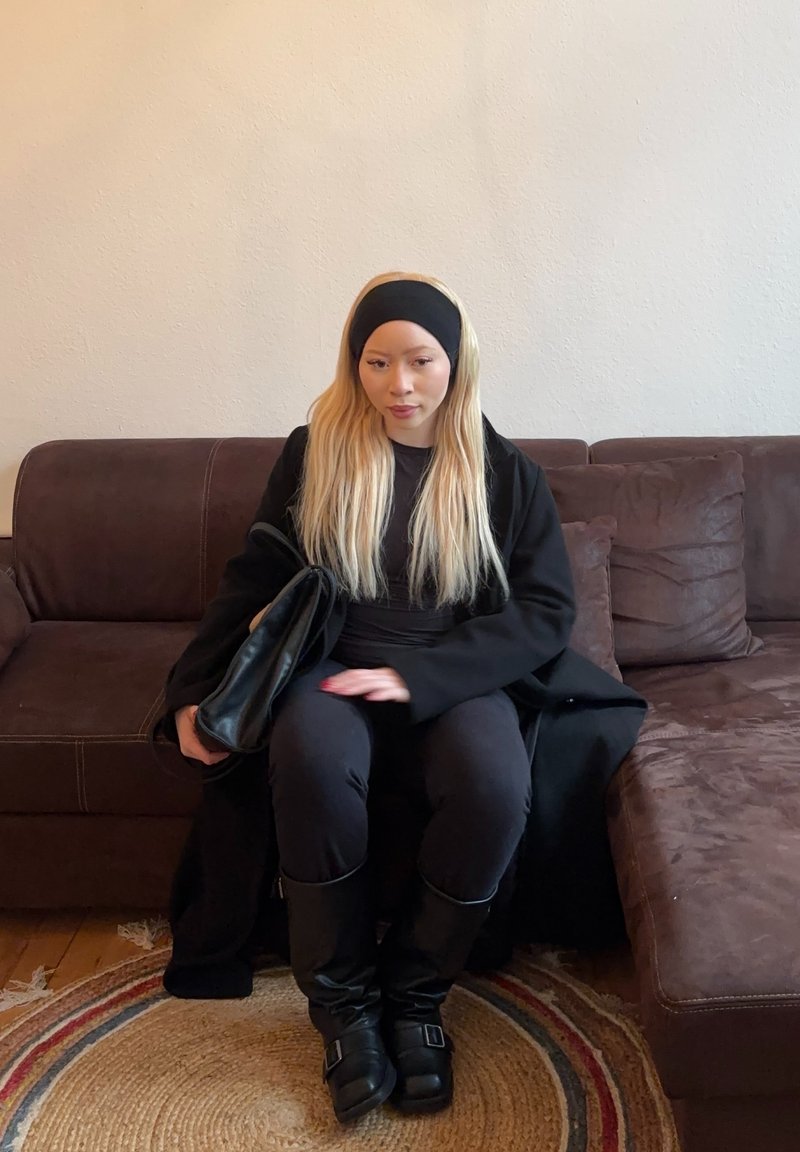 Black coat and top, black trousers, and knee-high black boots. Long blonde hair; seated on a dark brown sofa with a circular rug.