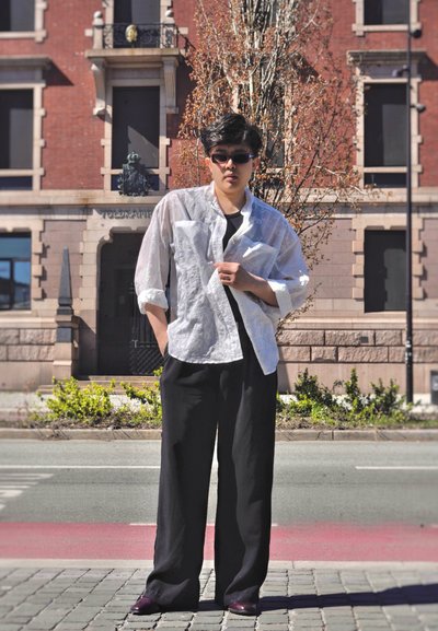 Persona con gafas de sol, camisa blanca semitransparente sobre top negro y pantalones negros holgados, de pie en la acera urbana cerca de la calle y un edificio de ladrillo.