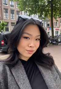 Manteau en laine gris avec une finition texturée, tenue noire en dessous et des lunettes de soleil reposant sur la tête, le tout se déroulant dans une scène de rue.