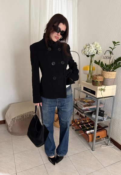 Mujer con chaqueta negra de doble botonadura, gafas de sol, vaqueros azules y zapatos negros, sostiene un bolso negro y está de pie junto a una estantería metálica con plantas y decoraciones.