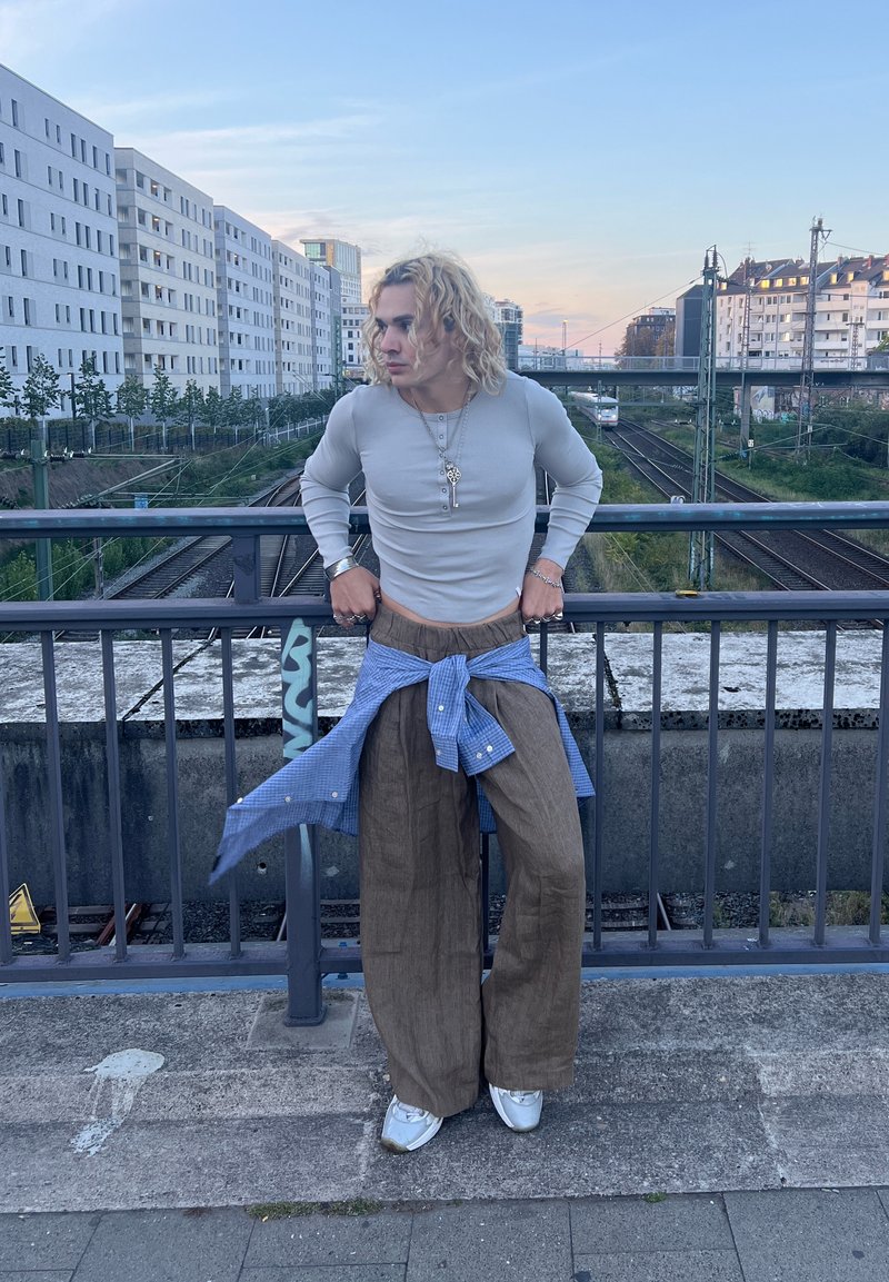 Lichtgrijze lange mouwen shirt, bruine wijde broek, blauwe geruite blouse om de taille geknoopt, witte sneakers. Achtergrond toont een stedelijke omgeving.