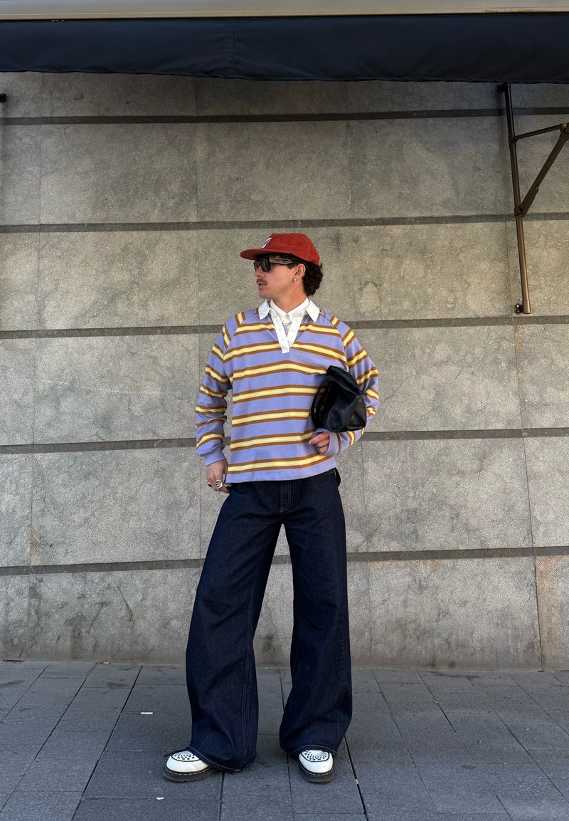 Personne portant une casquette rouge, une chemise rayée à manches longues, un jean large foncé, tenant une pochette noire, debout contre un mur en pierre.