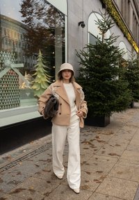 Béžový dvojradový vlnený kabát s výšivkou, krémový pletený sveter, biele nohavice so širokými nohavicami, béžové lodičky na podpätku a béžový klobúk typu bucket.