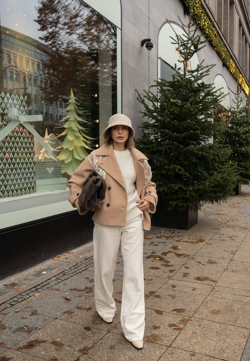 Casaco bege de lã com abotoamento duplo e bordados, camisola de malha creme, calças largas brancas, sapatos de salto bege e chapéu tipo balde bege.