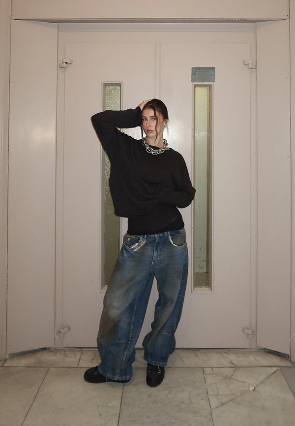 Tânără purtând un pulover negru supradimensionat, blugi albăstriți largi și un colier argintiu gros, pozează în fața unor uși duble bej închise.