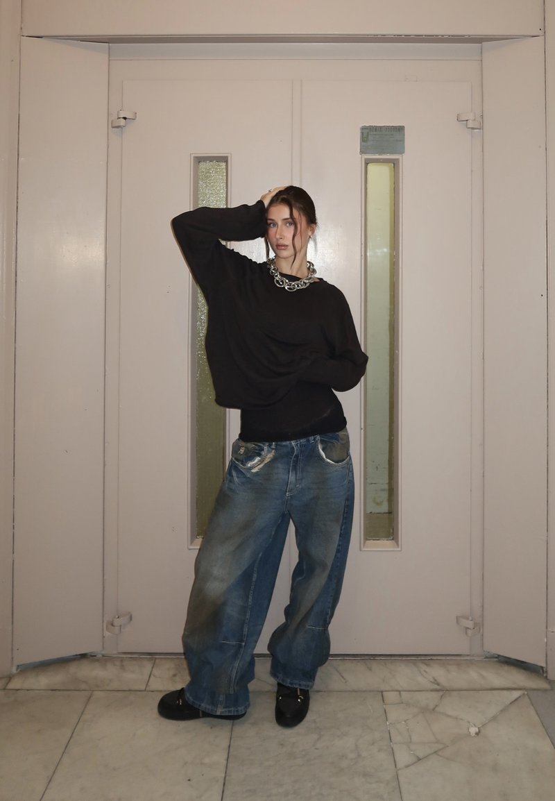 Nuori nainen, joka pukeutunut ylisuurtaan mustaan neuleeseen, leveisiin sinisiin farkkuihin ja rotevaan hopeiseen kaulakoruun, poseeraa suljettujen beigejen kaksoisovien edessä.