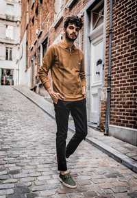 Langermet brun polo med glidelås og brystlomme, kombinert med svarte bukser og grønne sko, plassert på en brosteinsgate.