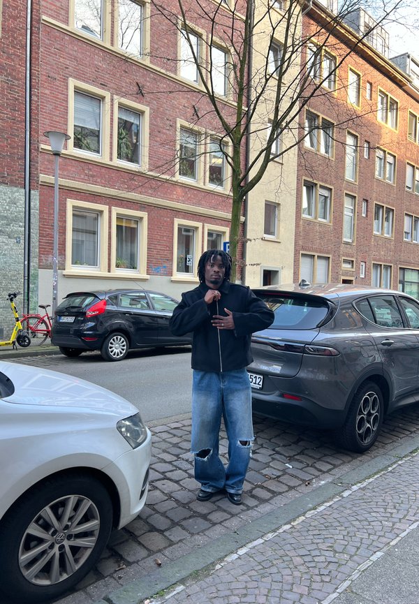 Uomo con dreadlocks che indossa una giacca nera e jeans strappati, in piedi su un marciapiede di ciottoli tra auto parcheggiate in una strada urbana.