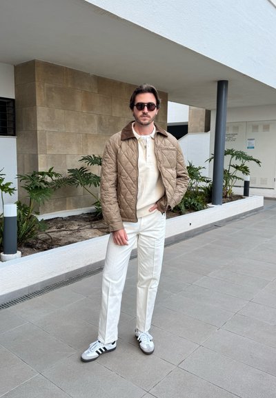 Chaqueta acolchada marrón con un cuello oscuro, suéter crema, pantalones blancos y zapatos grises con rayas negras. De pie frente a una pared de piedra.