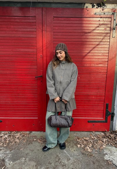 Joven mujer con chaqueta gris, sombrero marrón y pantalones verdes que sostiene un bolso marrón oscuro de pie frente a grandes puertas de madera rojas al aire libre.