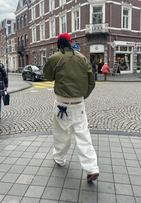 Personne avec des dreadlocks portant une casquette rouge, une veste verte, un pantalon blanc, marchant sur une rue pavée près d'une boutique de bagels dans une ville européenne.