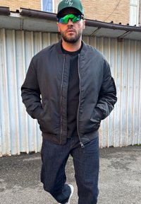 Fekete bomber dzseki bordázott mandzsettákkal, fekete ing fölött viselve, sötét farmerral és fehér tornacipővel párosítva; kiegészítők: zöld sapka és napszemüveg.