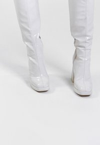 Frau in glänzenden weißen knielangen Plateauschuhen mit Blockabsätzen, die auf einer weißen Fläche steht.