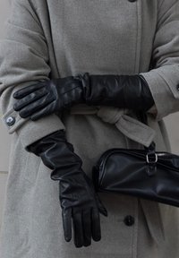 Černé kožené rukavice s přiléhavým střihem, s mírně naznačenou strukturou u zápěstí a hladkými konečky prstů, v kombinaci se šedým kabátem.