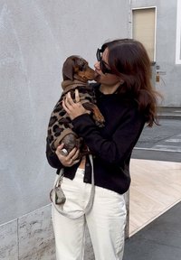 En brun dachshund i en beige leopardmønstret sweater holdes af en person iført en sort sweater og solbriller, med lyse bukser.