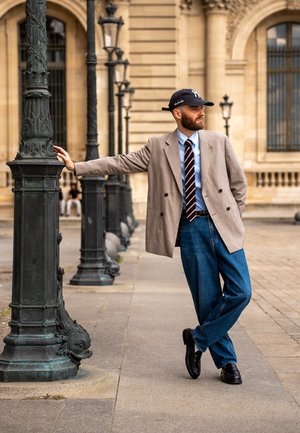 Hellgrauer, doppelt geknöpfter Blazer über einem blauen Hemd und einer weinrot gestreiften Krawatte, kombiniert mit weiten blauen Jeans und schwarzen Schuhen, stehend an einem Laternenpfahl.
