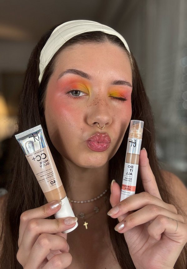 Joven con colorido maquillaje de ojos sostiene dos productos de IT Cosmetics, guiña un ojo y frunce los labios, lleva una cinta blanca en la cabeza y un collar con cruz.