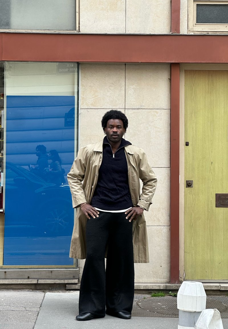 Man met een afro kapsel, gekleed in een beige trenchcoat, zwarte trui en wijde zwarte broek, staat met de handen in de zij op het stadsstoep.