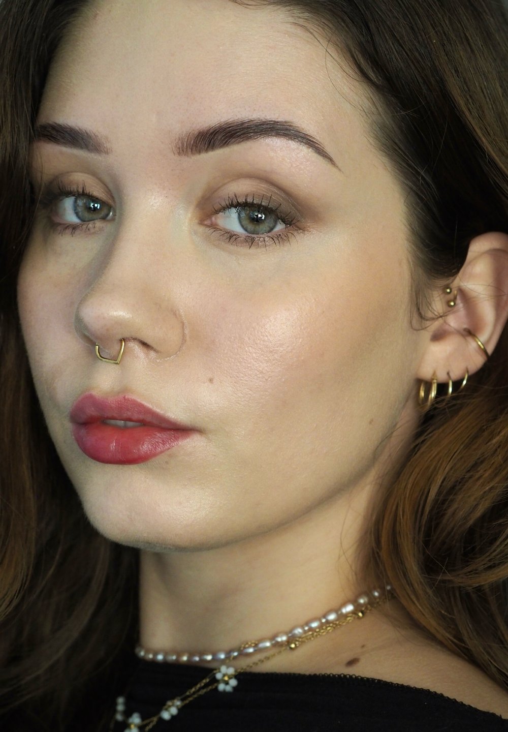 Close-up van een gezicht met een lichte huid, bruin haar en groene ogen. Opvallende kenmerken zijn een gouden septumring, verschillende gouden oorbellen en volle lippen met roze lippenstift.