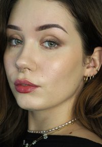 Close-up van een gezicht met een lichte huid, bruin haar en groene ogen. Opvallende kenmerken zijn een gouden septumring, verschillende gouden oorbellen en volle lippen met roze lippenstift.