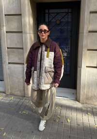 Femme portant une veste matelassée à blocs de couleur, un pantalon large beige, des baskets blanches et des lunettes de soleil foncées, tenant un sac à main à motif sur un trottoir en ville.