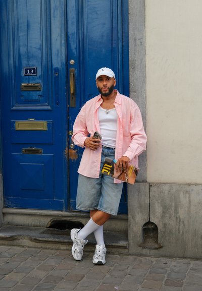 Camisa rosa claro, camiseta blanca sin mangas, pantalones cortos de mezclilla, calcetas blancas hasta la mitad de la pantorrilla y zapatillas plateadas. Sosteniendo una revista, apoyándose contra una puerta azul.