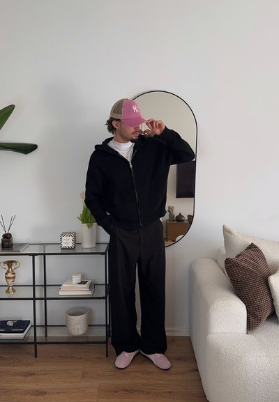 Sudadera de punto negra con cremallera, camiseta blanca, pantalones negros de pierna ancha y zapatillas rosas. Los accesorios incluyen una gorra rosa clara y joyas.