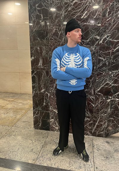 Suéter azul claro con un diseño esquelético blanco, combinado con pantalones negros y zapatillas con detalles verdes. Fondo de mármol.