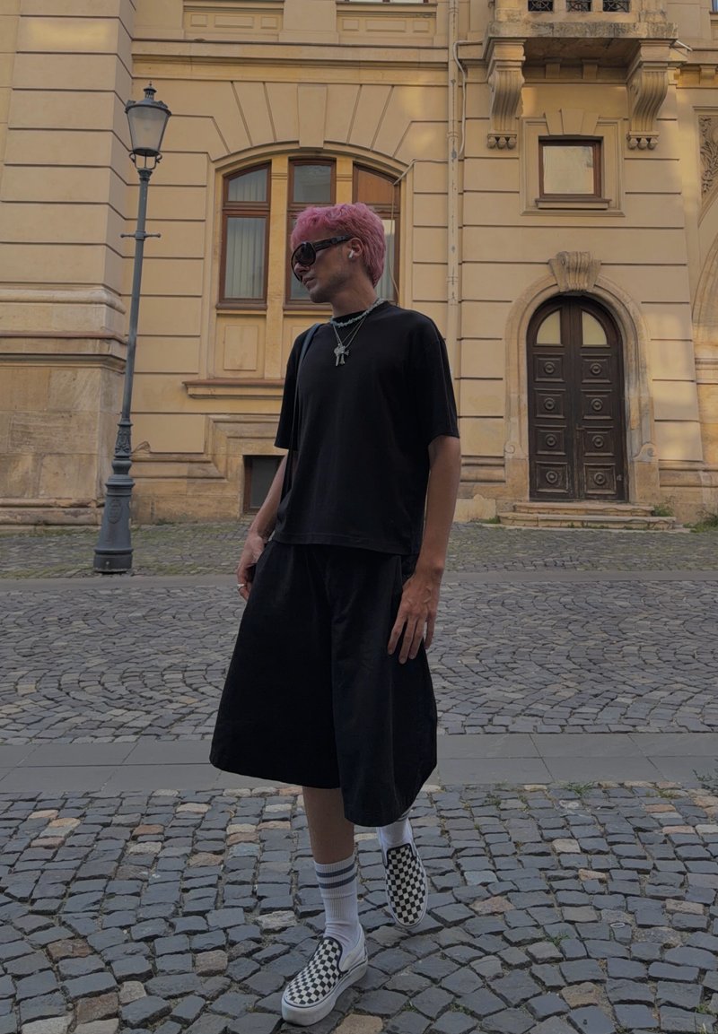 Henkilö, jolla on vaaleanpunaiset hiukset, pukeutuneena mustiin vaatteisiin, ruudullisiin slip-on-kenkiin ja aurinkolaseihin, seisoo mukulakivikadulla lähellä historiallista rakennusta.