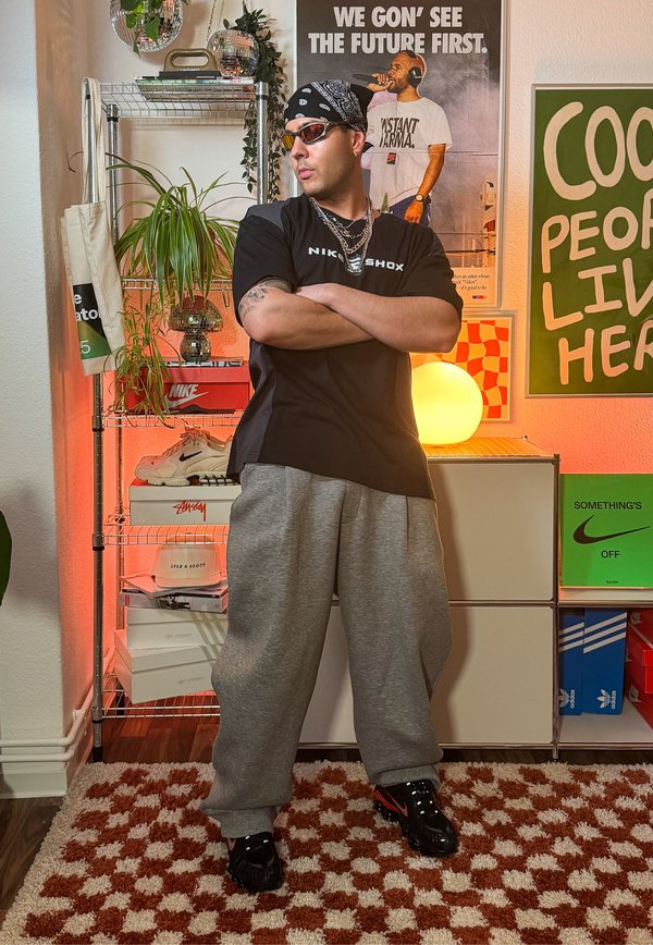 Svart Nike Shox t-shirt, grå sweatpants, svarta sneakers med röda detaljer. Accessoarer inkluderar solglasögon och en bandana, med en bakgrund av hyllor.