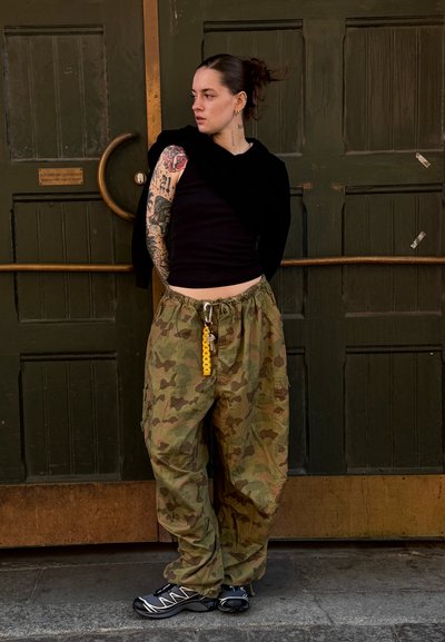 Pantalones cargo verde camuflaje con bolsillos, camiseta sin mangas negra y un llavero amarillo. De pie frente a una puerta de madera verde.