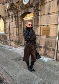 Veste en cuir marron à col haut, pantalon large assorti et bottes pointues montant jusqu'aux genoux. Sac à main noir et lunettes de soleil. Texture lisse.