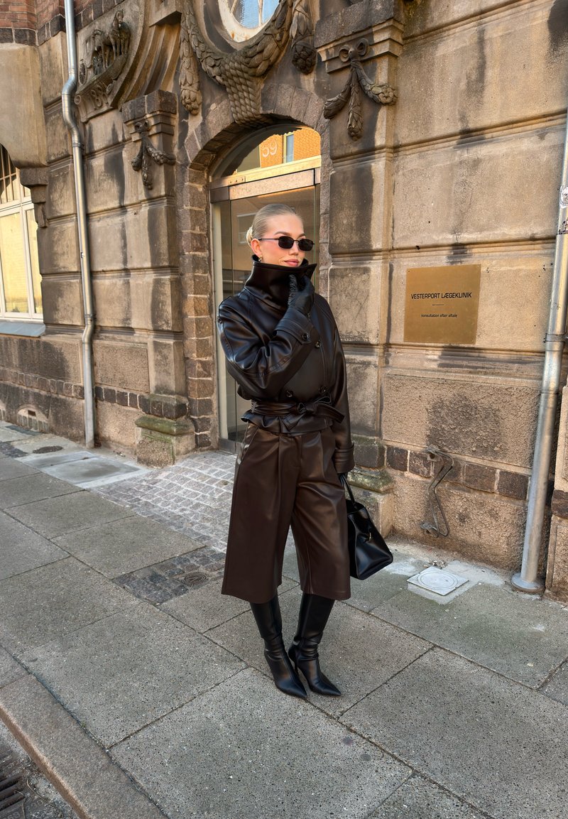 Veste en cuir marron à col haut, pantalon large assorti et bottes pointues montant jusqu'aux genoux. Sac à main noir et lunettes de soleil. Texture lisse.