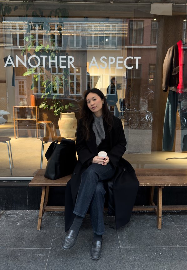 Cappotto nero, sciarpa grigia e denim scuro seduti su una panca di legno con una borsa nera, tengono una tazza di caffè davanti a una vetrina del negozio.
