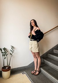 Črna denimska jakna, olivno zelena majica, bež kratke hlače, rjavi sandali; stoji ob lončnici na stopnicah s sivim granitnim robom.