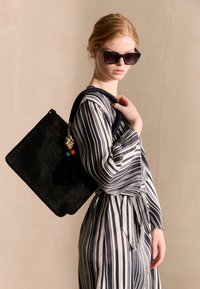 Zwarte geweven draagtas met een gestructureerde vorm, versierd met kleurrijke kralen en een toef. Model draagt een zwart-wit gestreepte jurk.