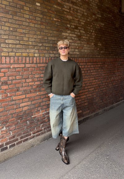 Persona con cabello rubio y gafas de sol se apoya contra una pared de ladrillos, vistiendo un suéter oliva, pantalones vaqueros anchos y desteñidos y botas de vaquero marrones.