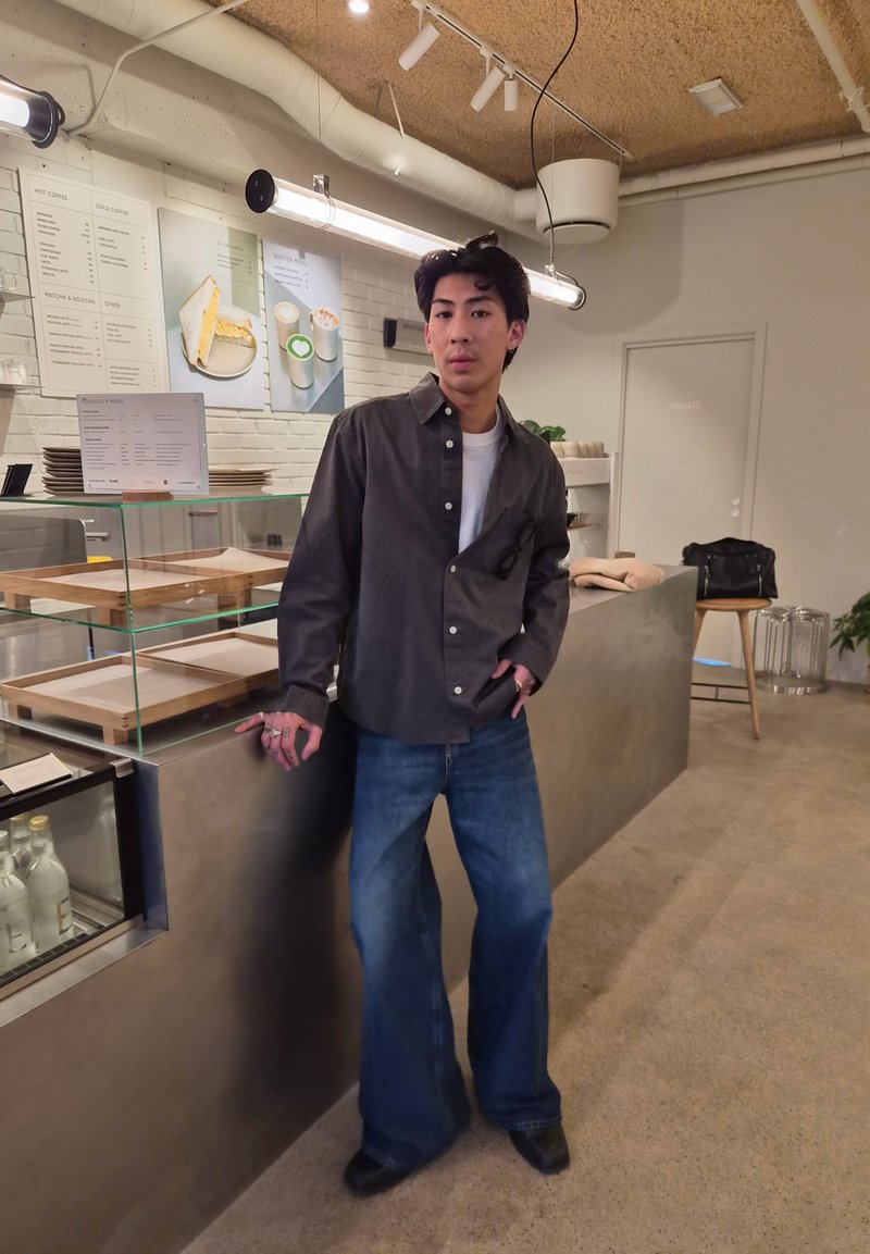 Jeune homme avec chemise foncée et jean large appuyé sur le comptoir du café avec vitrine en verre et menu au mur derrière lui.