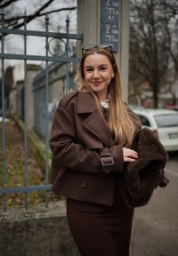 Νεαρή γυναίκα με μακριά ξανθά μαλλιά, φορώντας καφέ παλτό και φούστα, κρατώντας μια καφέ γούνινη τσάντα, στέκεται δίπλα σε μεταλλικό φράχτη σε μια νεφελώδη μέρα.