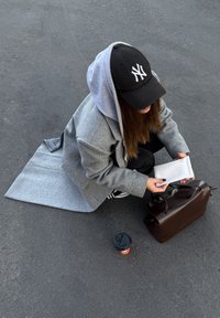 Manteau en laine grise superposé sur un hoodie gris clair, casquette noire, tenant un portefeuille blanc, avec un sac en cuir marron et une tasse de café sur le sol.