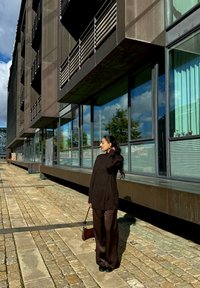 Pullover marrone oversize e pantaloni a gamba larga, con finitura testurizzata. Il modello porta una borsa marrone, in piedi di fronte a un'architettura moderna in vetro.