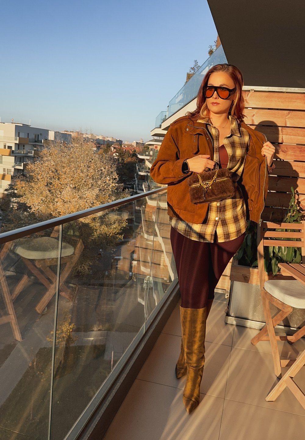 Giacca in camoscio marrone sopra una camicia a quadri, abbinata a leggings bordeaux e stivali gialli alti fino al ginocchio con trama. È presente una piccola borsa di pelliccia.